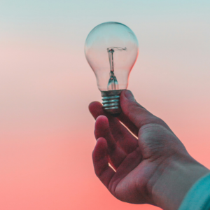 A hand holding a lightbulb in front of a dawn-colored horizon.