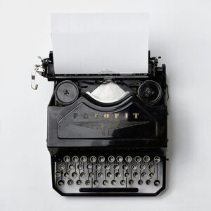 Black typewriter on white background looked at from above. A sheet of paper reading "*** The revolution will not be monospaced ***" is drawn in, the ribbon and brand "Favorit" together with a logo are visible as well as all of the keys (numbers and letters). Each key is round with a golden letter against a black surface. It is a QWERTZ-keyboard.