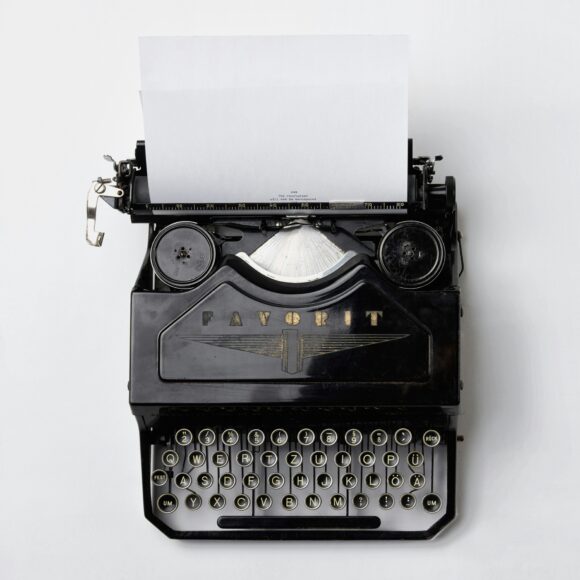 Black typewriter on white background looked at from above. A sheet of paper reading "*** The revolution will not be monospaced ***" is drawn in, the ribbon and brand "Favorit" together with a logo are visible as well as all of the keys (numbers and letters). Each key is round with a golden letter against a black surface. It is a QWERTZ-keyboard.
